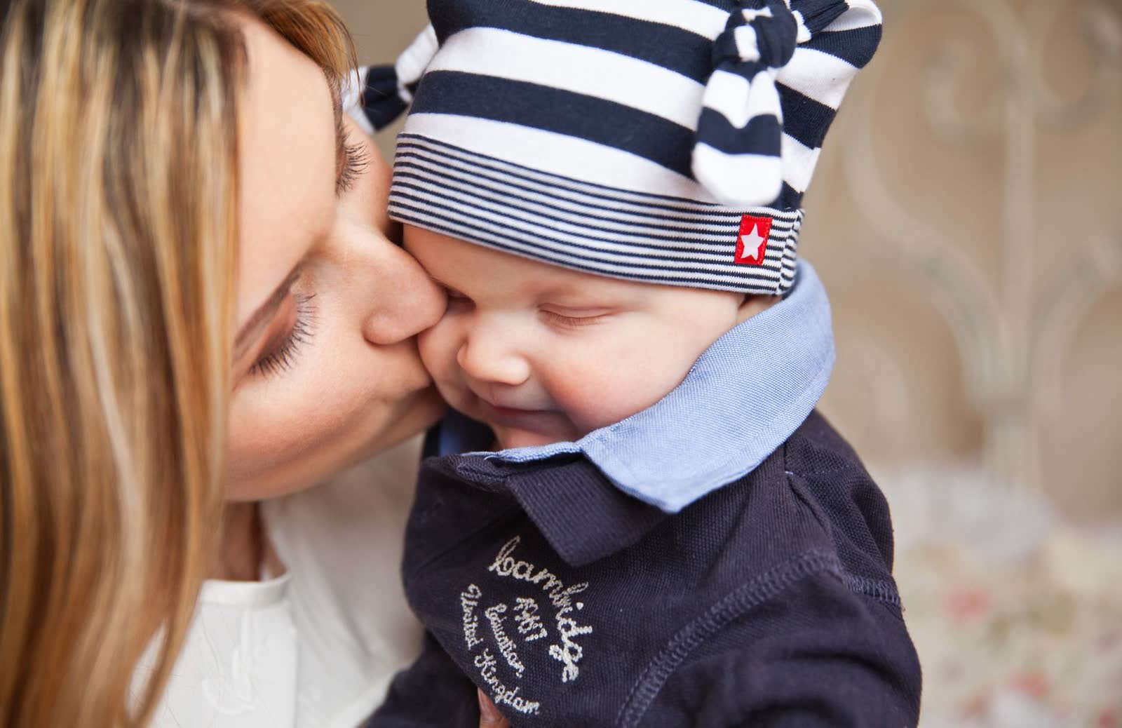 Mãe beijando a bochecha de um bebê com gorro listrado, que sorri de olhos fechados, em close.