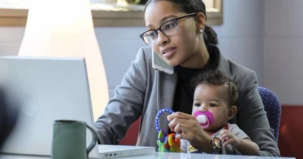Mulher de blazer e óculos falando ao telefone na mesa de trabalho, com o bebê de chupeta no colo